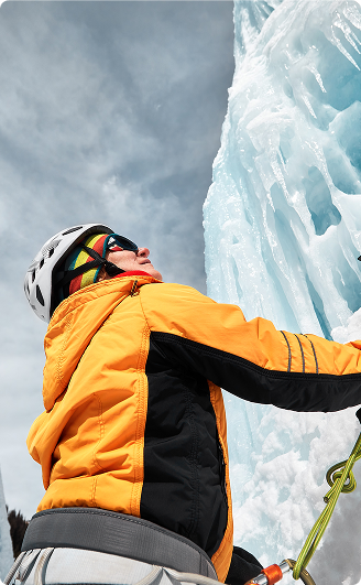 Ice climber in a yellow and black jacket looking up while ascending a frozen waterfall with climbing gear